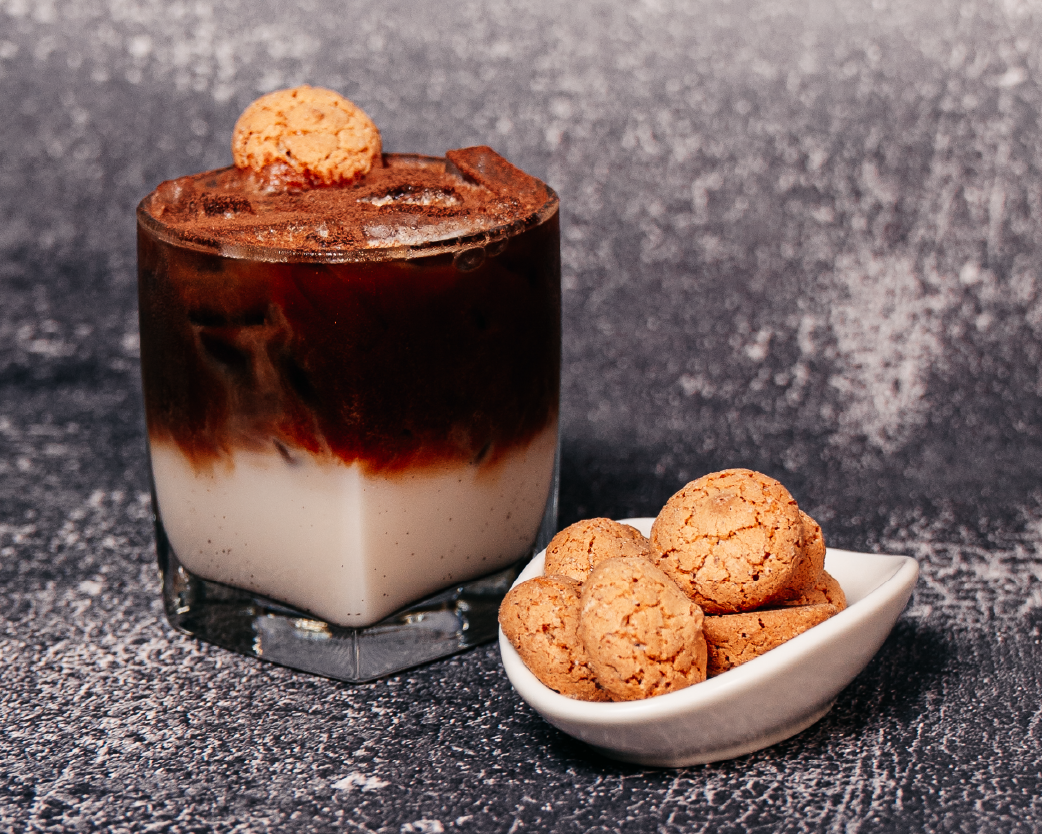 Layered iced espresso drink in a clear glass, topped with a cookie and styled with almond cookies on a textured surface.