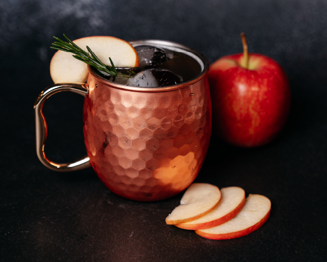 Copper mug filled with apple cider mocktail, garnished with apple slices and herbs on a dark background.