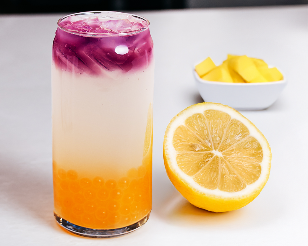 Layered iced beverage with purple, cream, and orange hues topped with ice, served alongside a halved lemon and mango pieces.