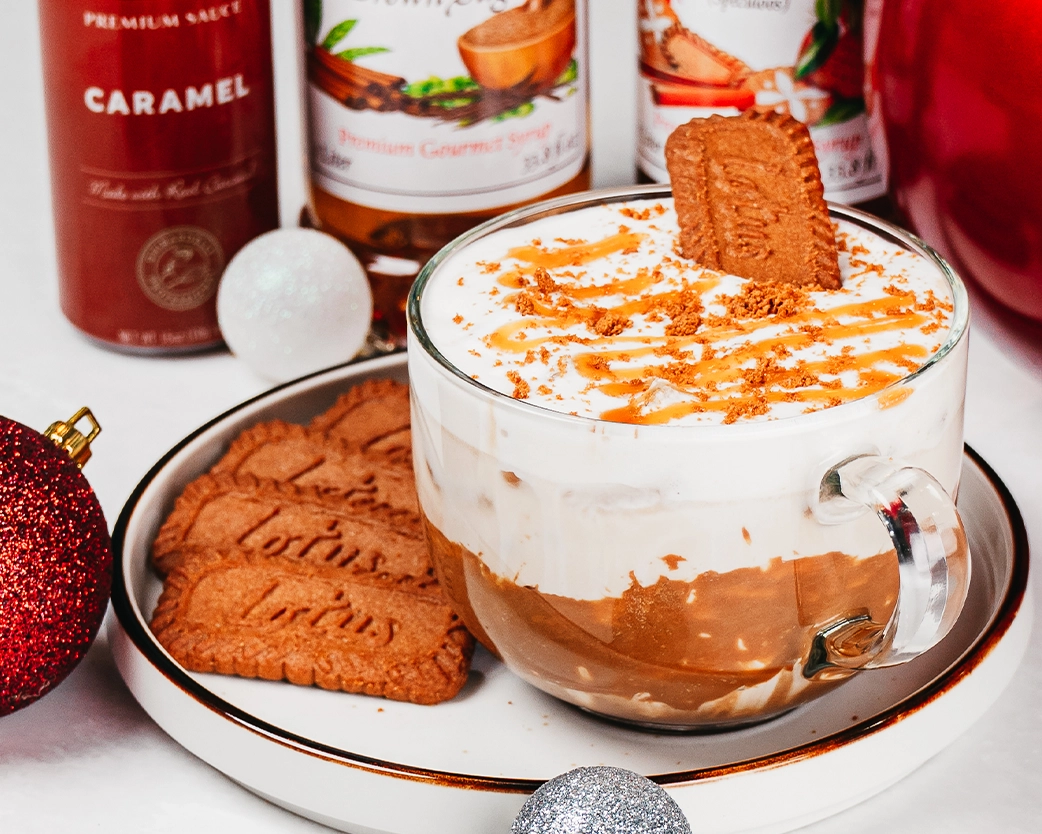 Iced latte in a clear glass mug topped with whipped foam, caramel  drizzle, and a biscuit, styled with cookies and syrup bottles.