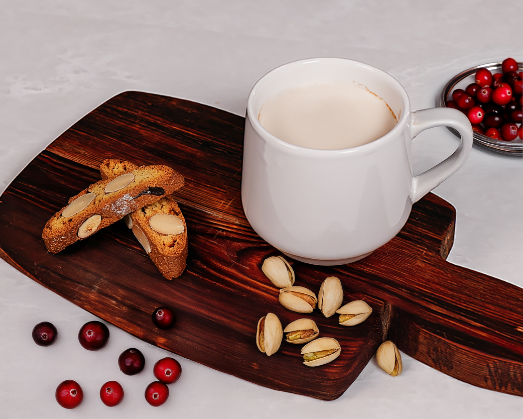 Hot latte in a white mug on a wooden board, styled with biscotti, pistachios, and fresh cranberries.