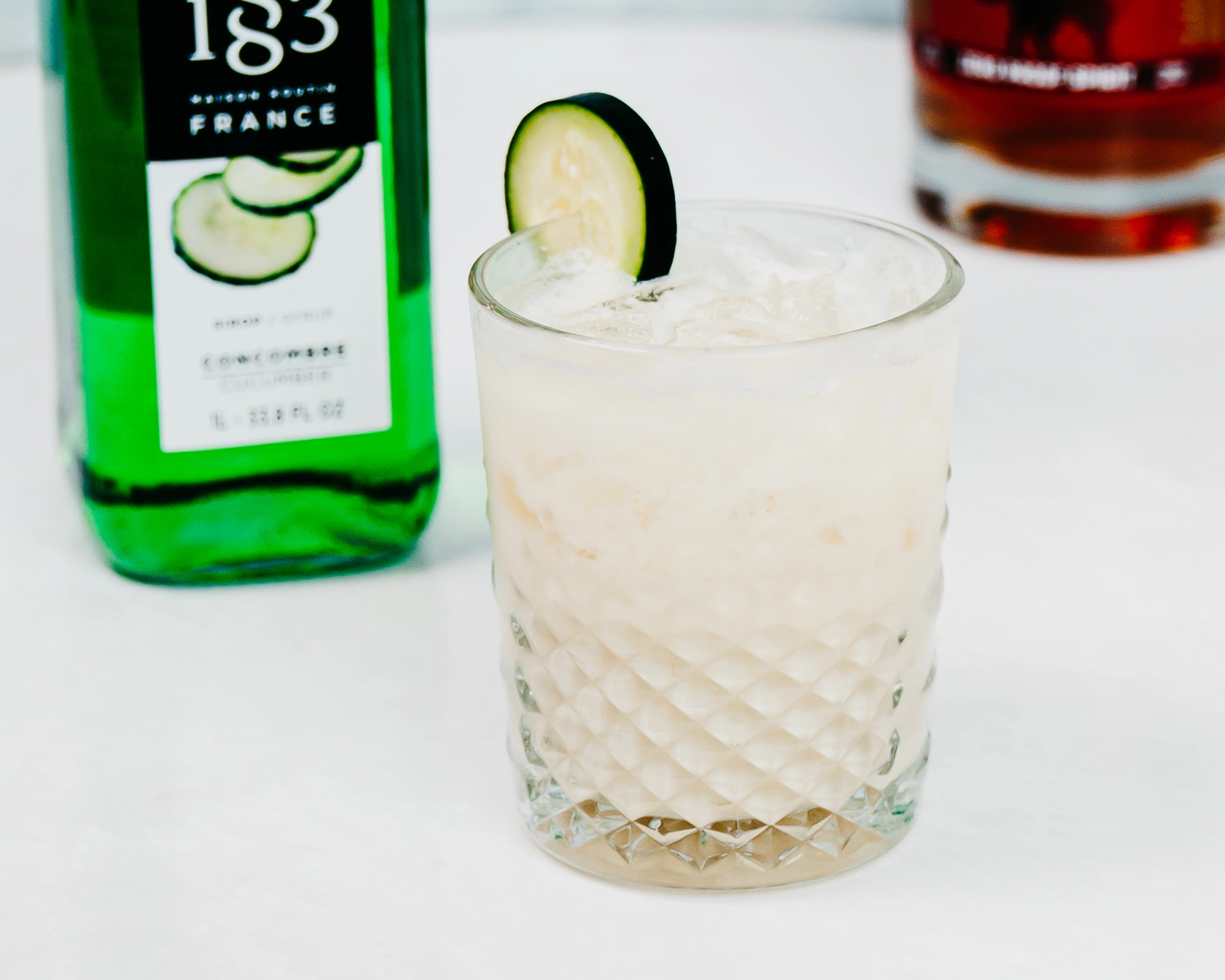 A creamy mocktail served in a short crystalized glass, garnished with a cucumber slice.