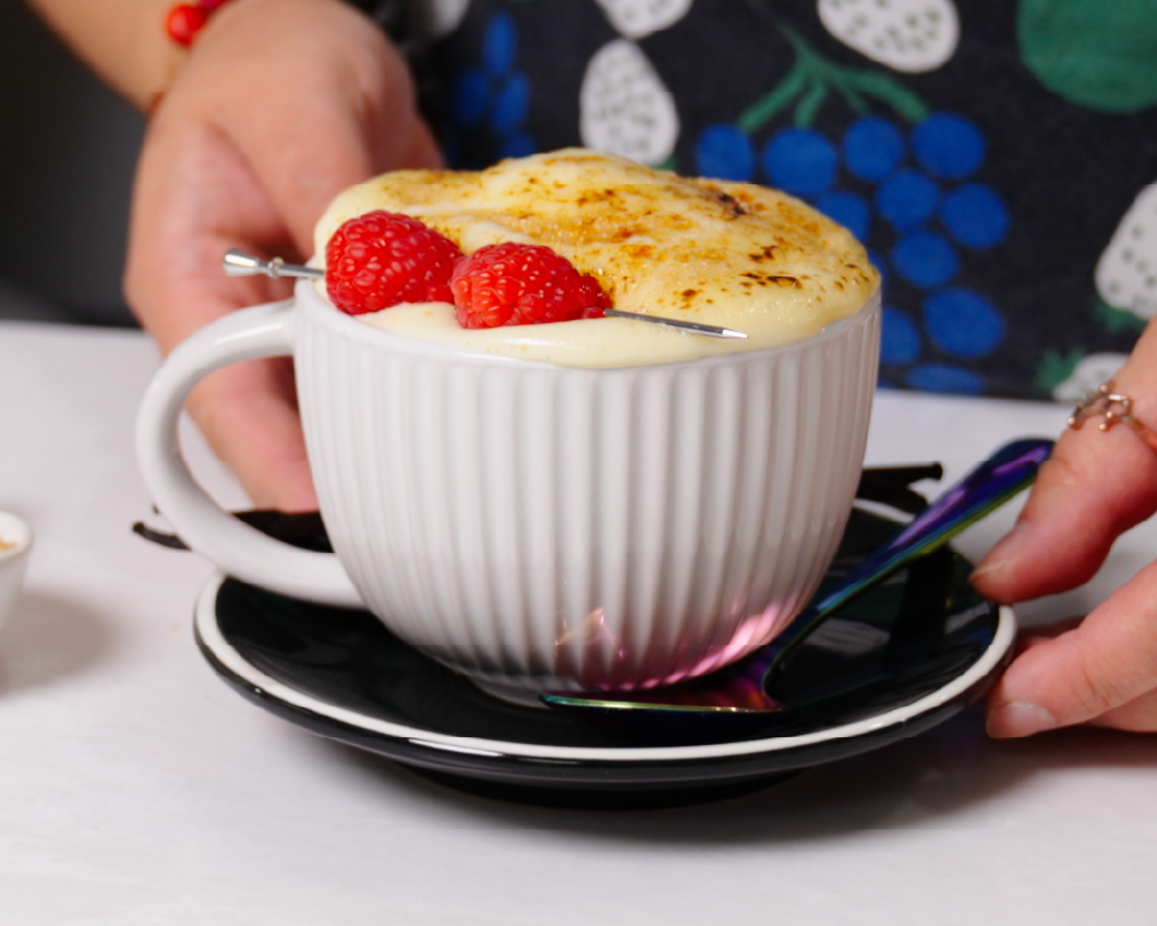 Hot cappuccino in a white cup with a torched custard-style topping and fresh raspberries, served on a black saucer.