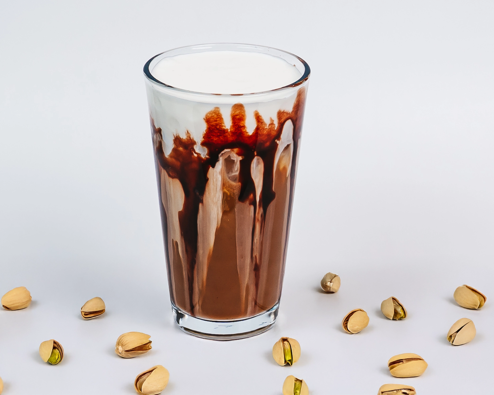 Iced mocha latte in a clear glass with chocolate syrup drizzled along the sides, creating a bold marbled effect of milk and cocoa. Pistachios scattered on a clean, neutral background.