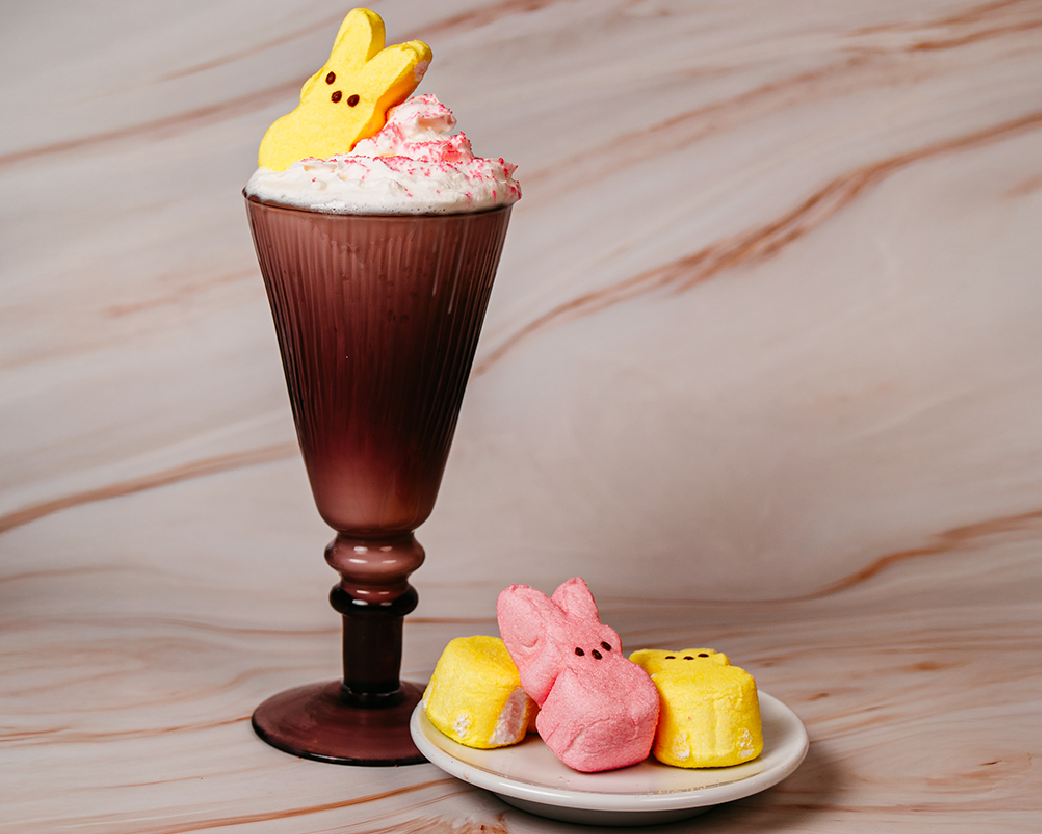 Frappe beverage topped with whipped cream and a bunny-shaped marshmallow, served in a tall glass with pastel marshmallow candies on the side.
