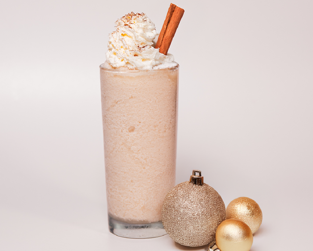 Creamy blended drink in a tall glass topped with whipped cream and a cinnamon stick, styled with gold ornaments.