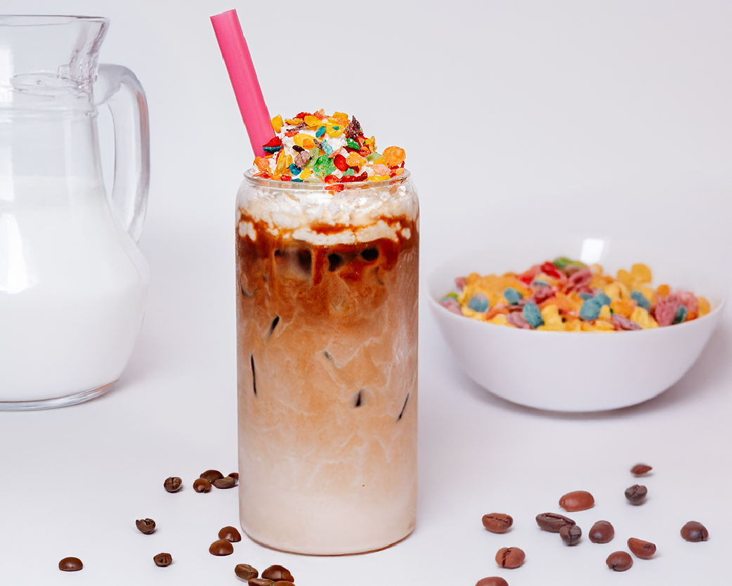 Layered iced coffee topped with whipped cream and colorful cereal pieces, styled with a milk pitcher and cereal bowl.