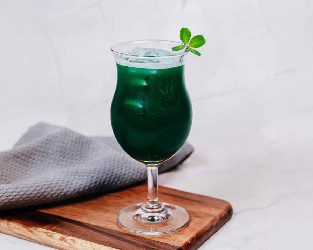 Deep green iced beverage in a stemmed glass, garnished with a clover leaf on a wooden board.