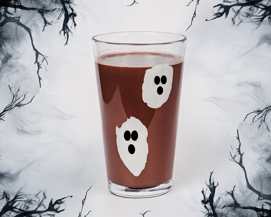 Iced chocolate drink in a clear glass decorated with ghost shapes, styled against a spooky illustrated background.