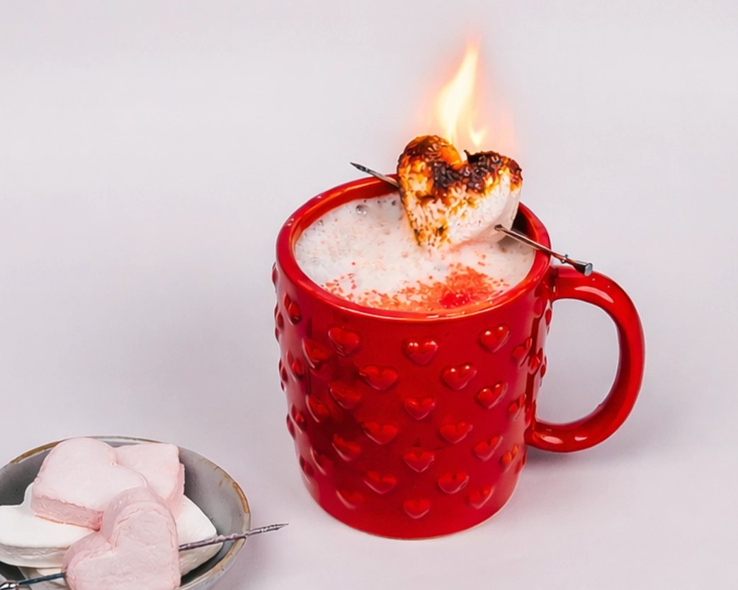 Flaming heart-shaped marshmallow perched on a creamy latte in a red heart-embossed mug, styled for Valentine’s Day.