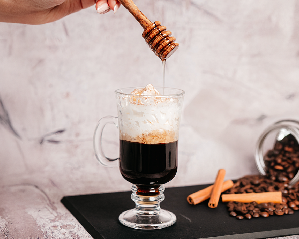 Hot coffee in a clear glass mug topped with foam, with honey being drizzled from a wooden dipper above.