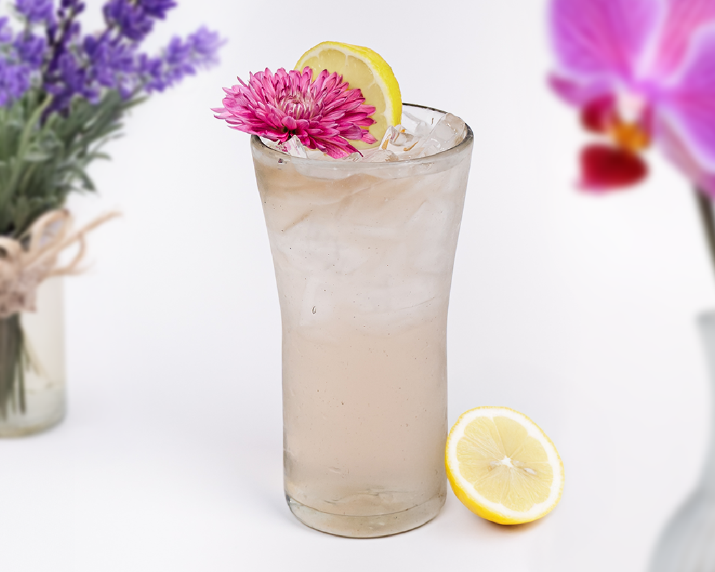 Light iced lemon beverage in a tall glass, garnished with a pink flower and lemon slice against a bright background.