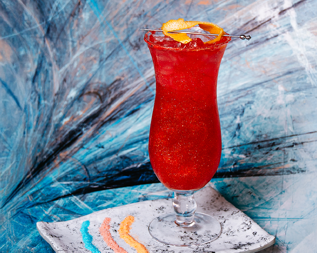 Bright red iced beverage in a curved glass, styled with colorful candy and an abstract blue background.