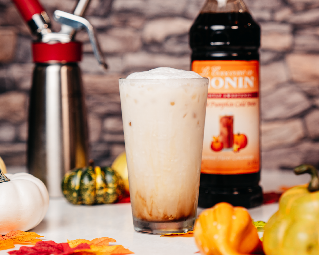 Layered cold brew topped with creamy foam, styled with pumpkins and syrup bottle in the background.