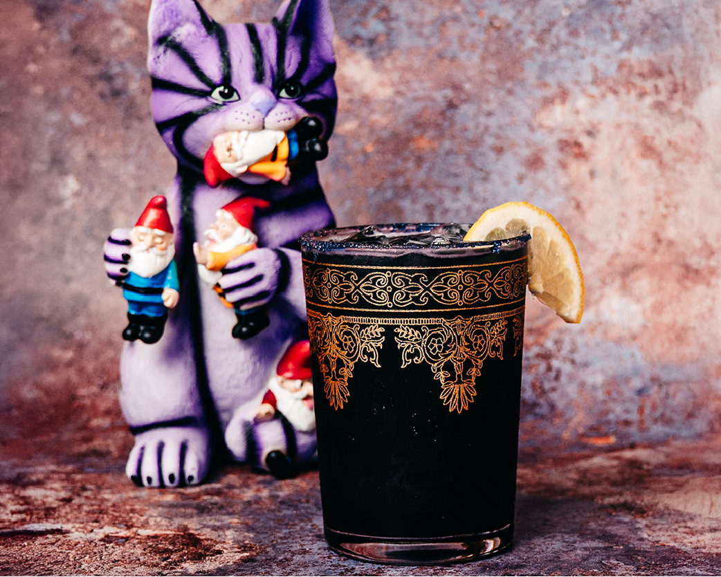 Dark iced beverage in a decorative black cup, styled with a whimsical monster figurine and moody backdrop.