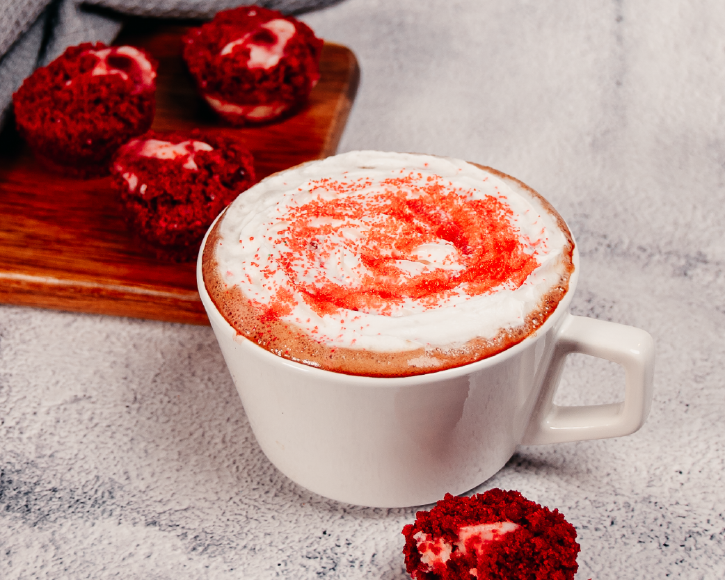 Hot latte in a white mug topped with foam and red velvet crumb garnish, styled with cake pieces.