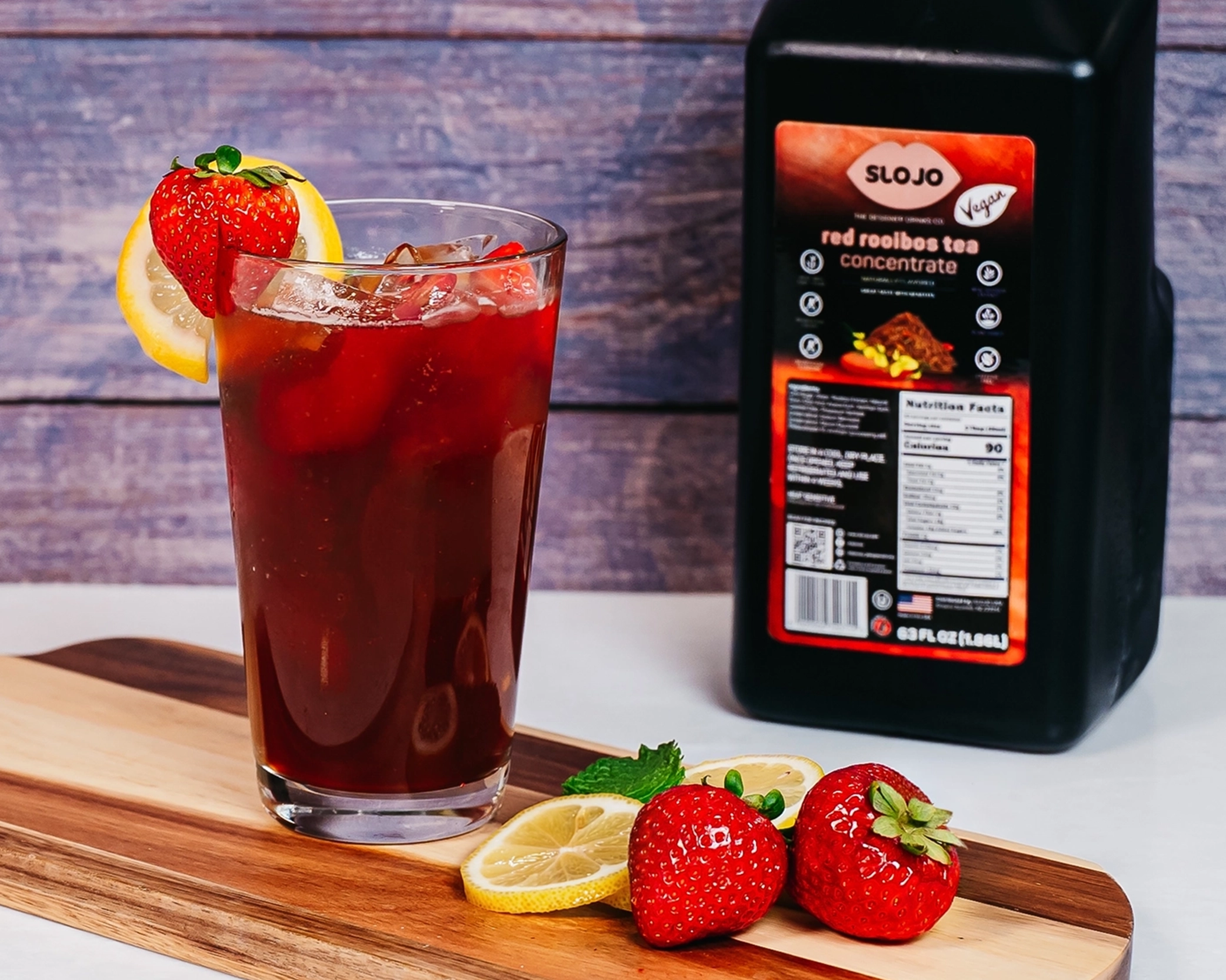 Iced red rooibos tea garnished with fresh strawberries and lemon slices, served beside a bottle of SloJo Red Rooibos Tea Concentrate.