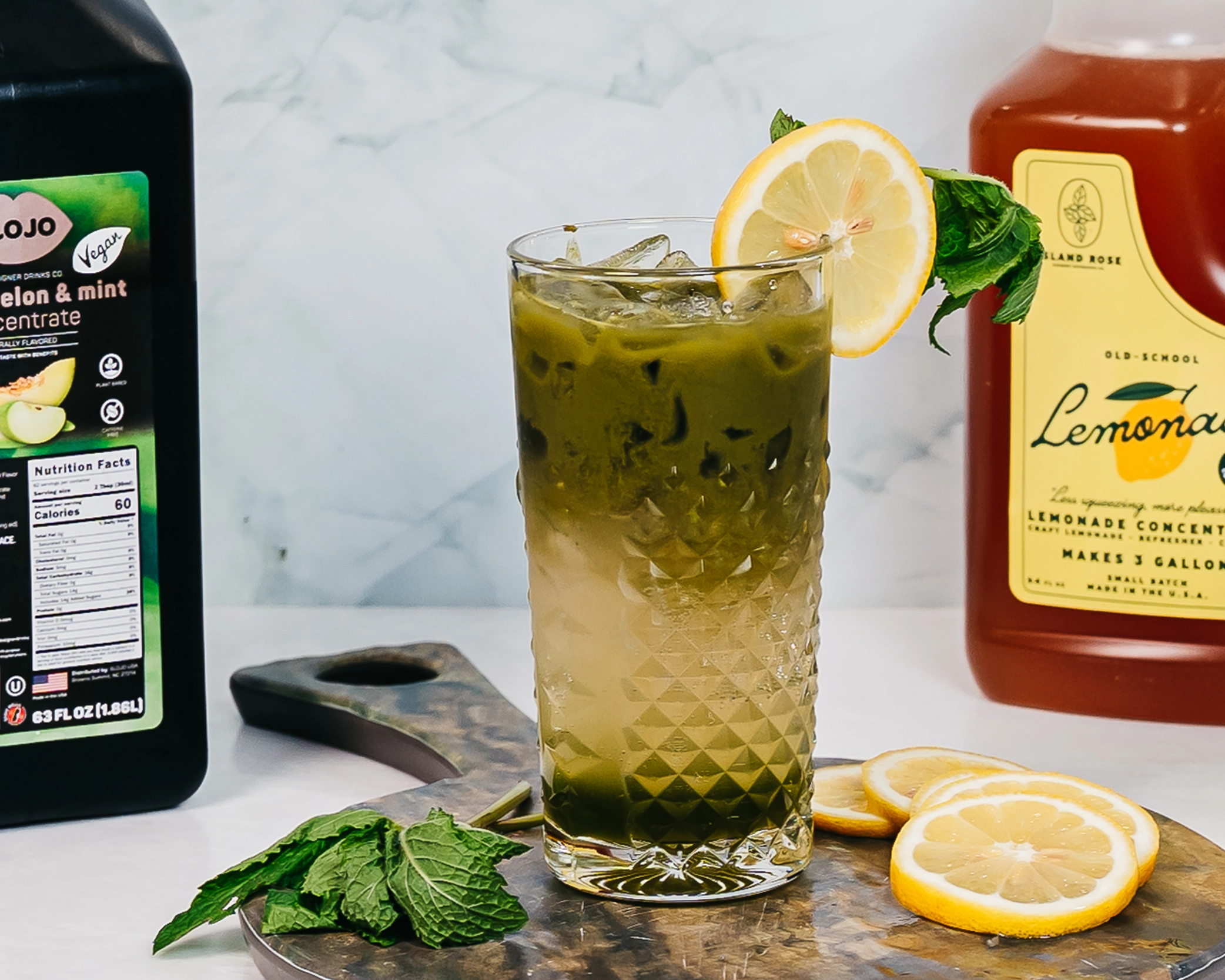 Layered iced lemonade garnished with fresh lemon and mint, served beside SloJo Apple, Melon, and Mint Concentrate and Island Rose Lemonade Concentrate.