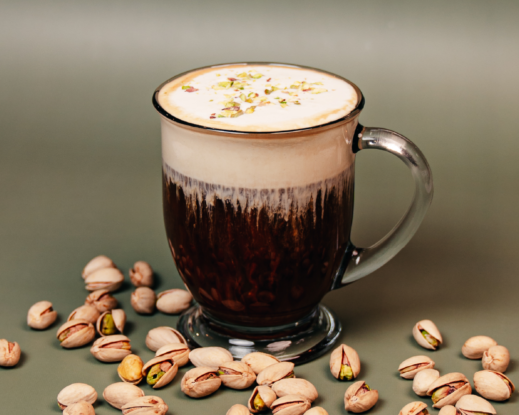 Layered pistachio iced coffee in a clear glass mug, topped with creamy foam and crushed pistachios, surrounded by whole pistachio nuts.