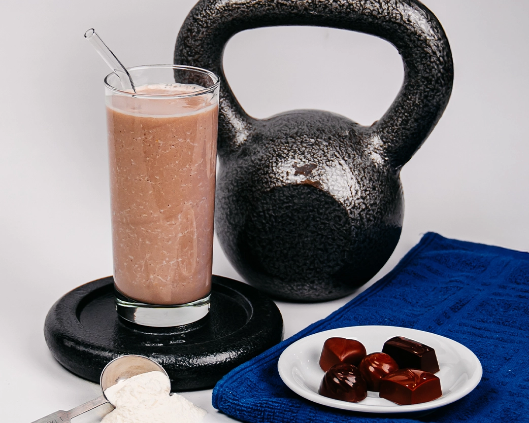 Chocolate protein shake in a tall glass with a straw, styled with a kettlebell weight, chocolate pieces, and a blue towel.