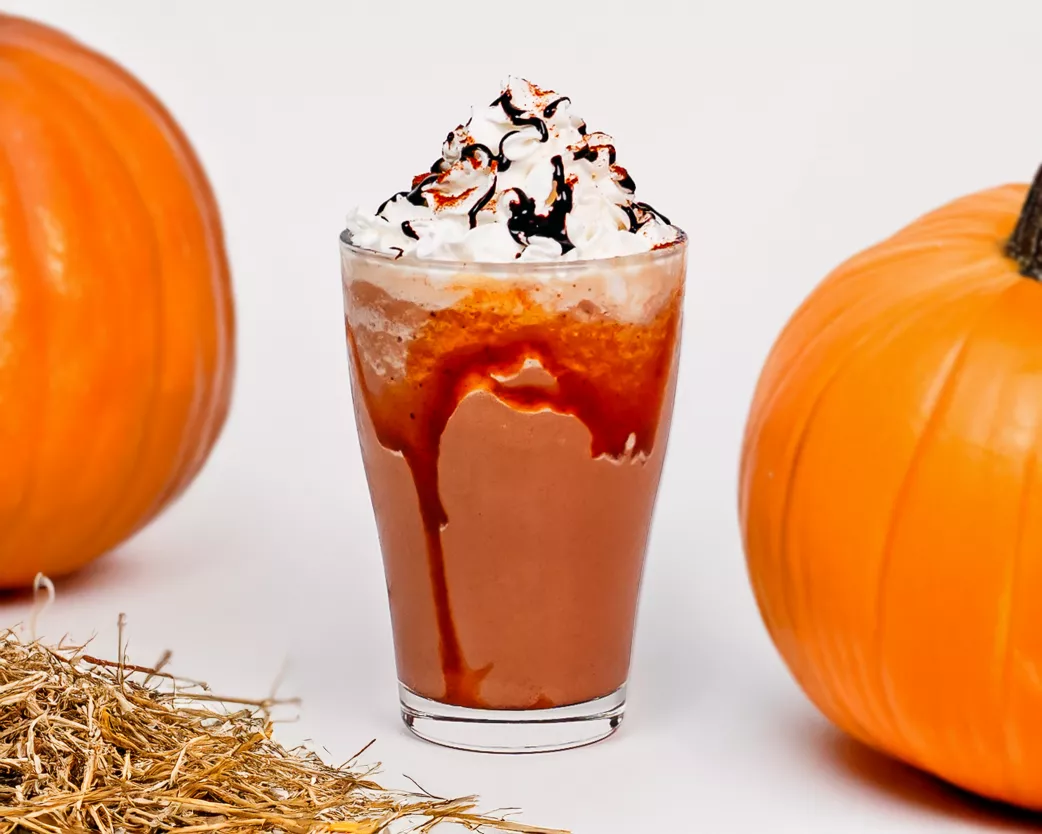 Blended fall-inspired frappe topped with whipped cream and drizzle, styled with pumpkins and straw accents.