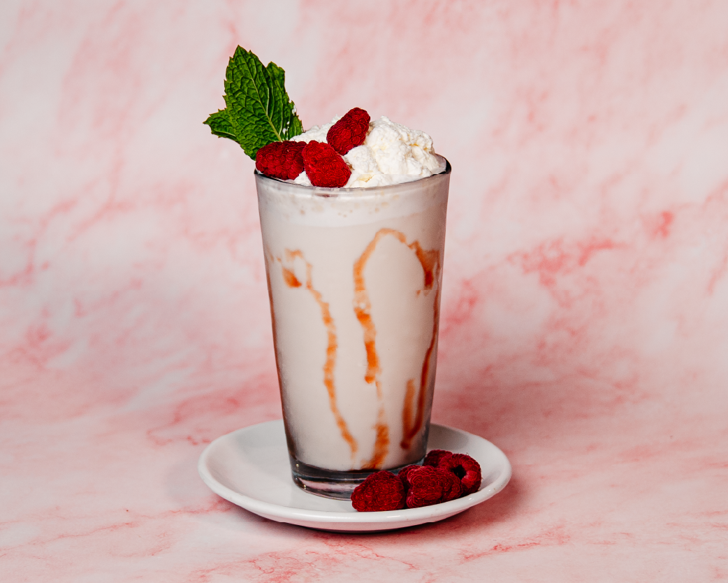 Creamy blended frappe drizzled with white chocolate sauce, topped with whipped cream, fresh raspberries, and mint in a tall glass.