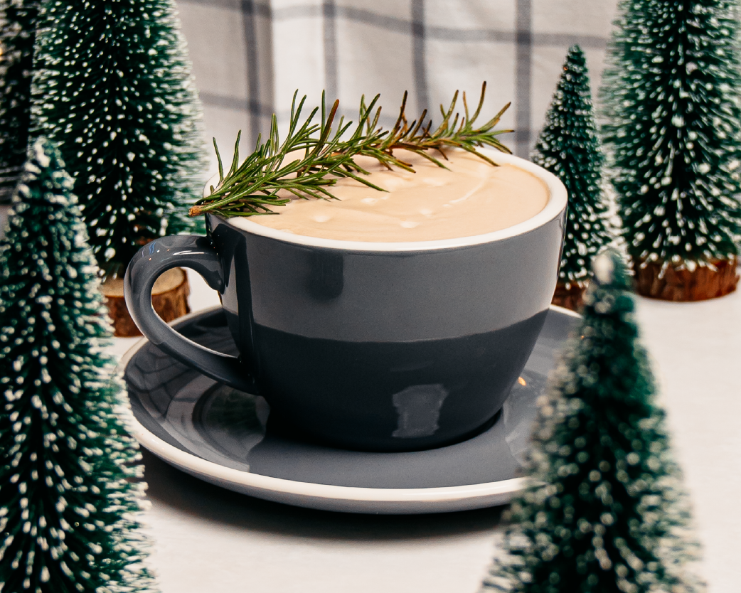 Creamy hot latte in a dark cup, garnished with a rosemary sprig and surrounded by miniature evergreen trees.