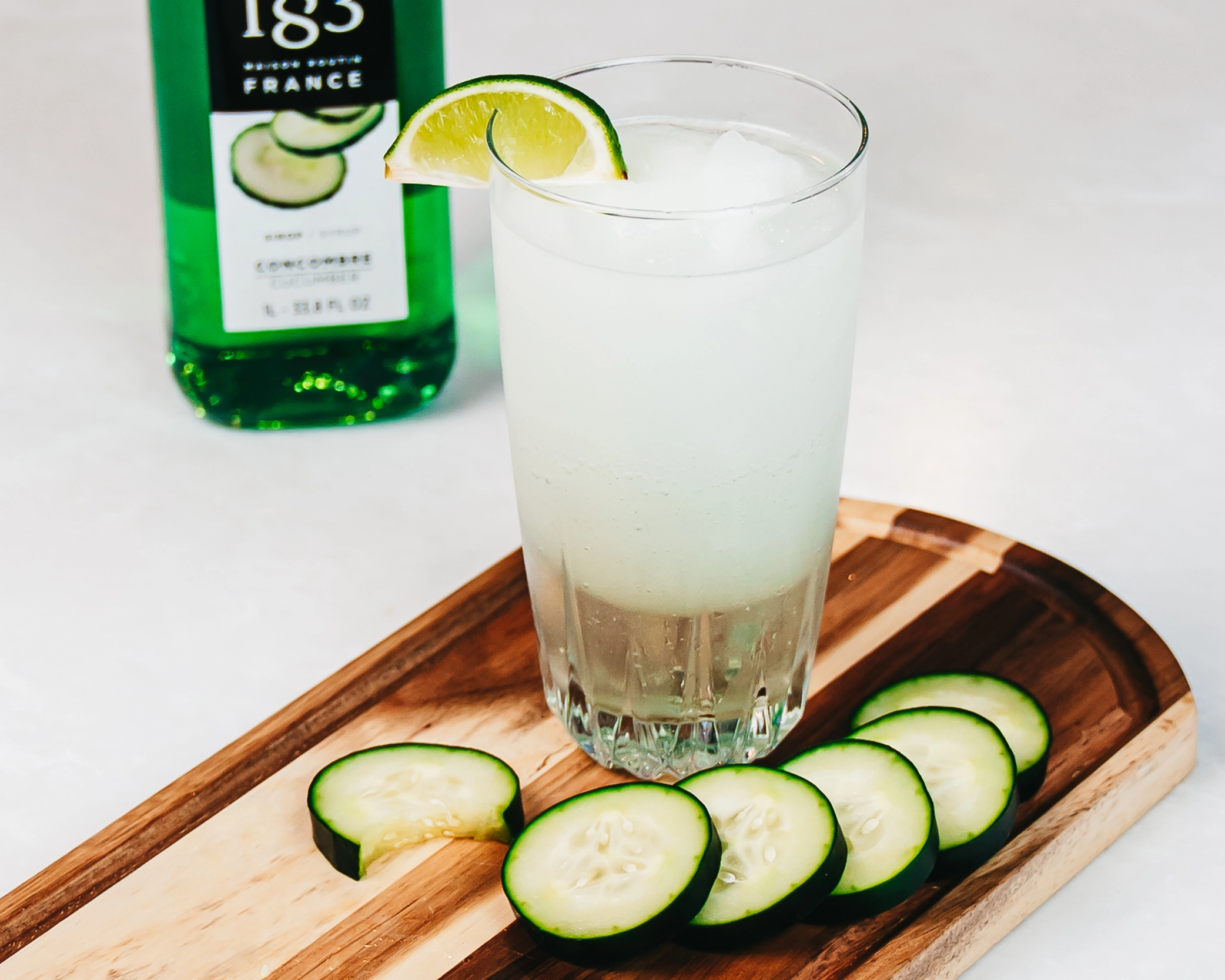 A blended iced beverage in a pint glass served with slices of cucumbers.